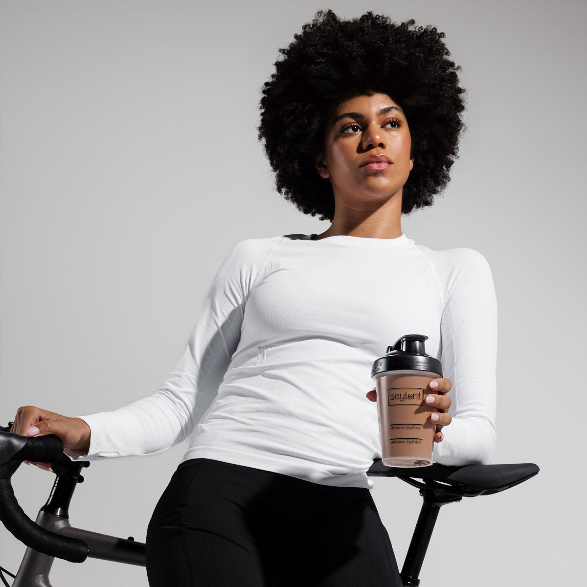 Woman holding Soylent chocolate complete protein shake while standing next to bike.